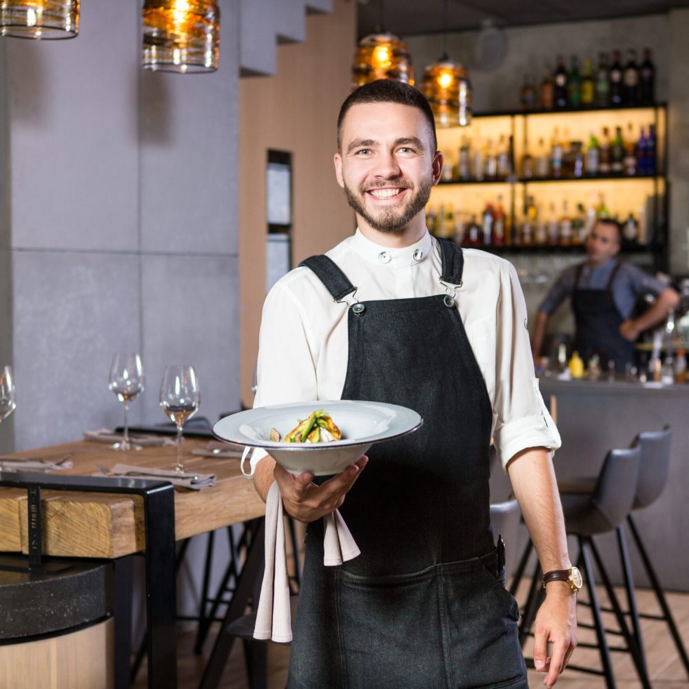 A handsome young guy with a beard dressed in an apron standing in a restaurant and holding a white plate with a moth. Against the background, the bar counter and the loft style interior.